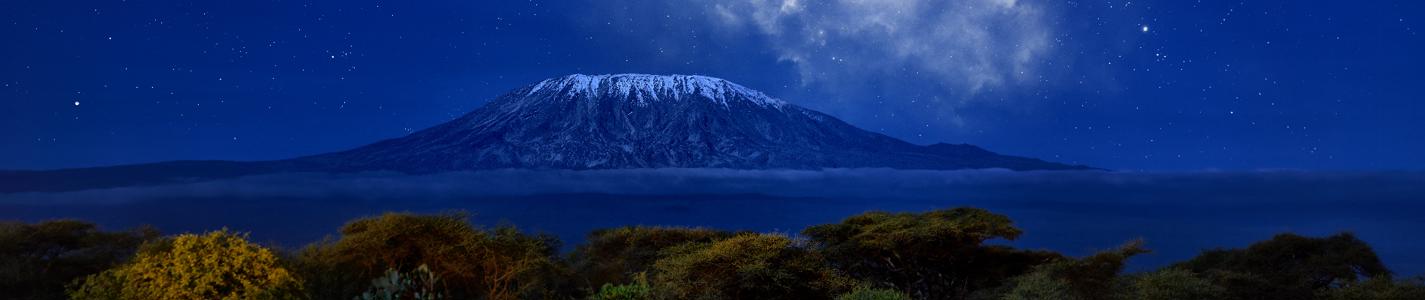 Stars over Mount Kilimajaro. Panoramic, night scenery of snow capped highest african mountain, lit by full moon against deep blue night sky with stars. Savanna view, Amboseli national park, Kenya. 